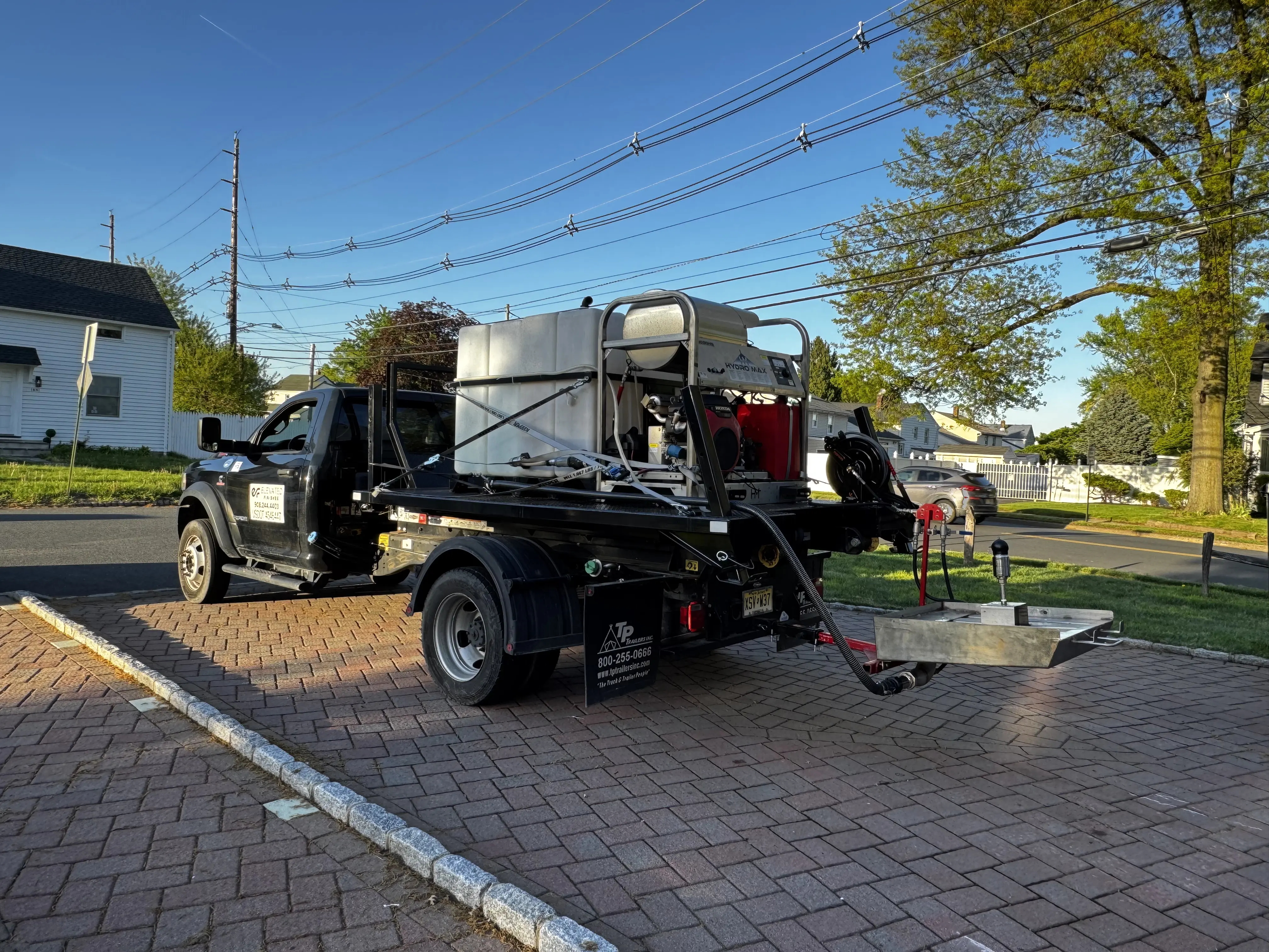 A black flatbed work truck parked on a brick driveway in a suburban neighborhood during sunny weather. The truck carries industrial equipment including a large water tank, hoses, and a pressure-washing or utility system mounted on the back. Trees, houses, and overhead power lines are visible in the background under a clear blue sky.