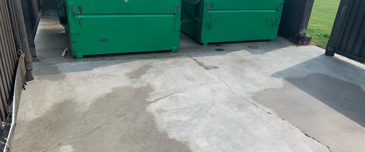 A concrete slab with two large green dumpsters and a dark fence visible on either side.