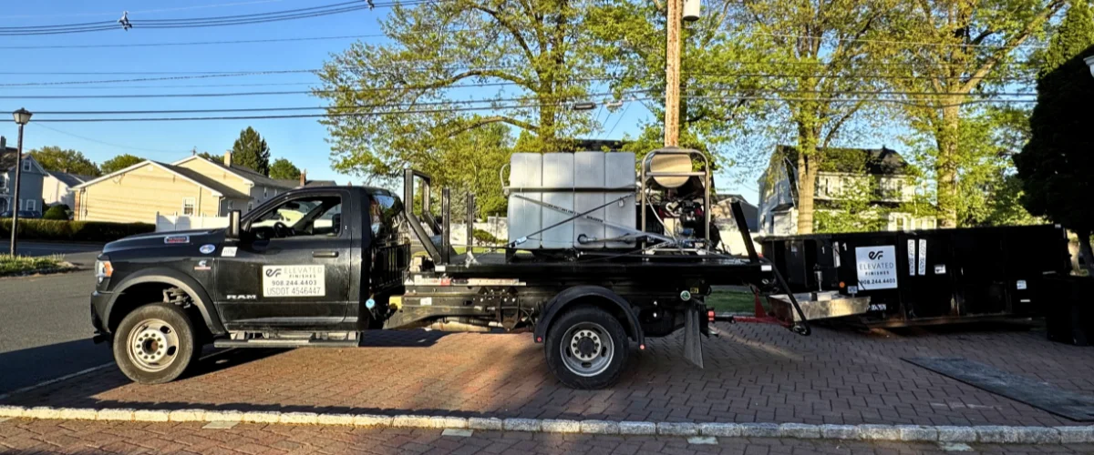 A black Ram truck with a flatbed and water tank, towing a black dumpster trailer on a brick driveway.