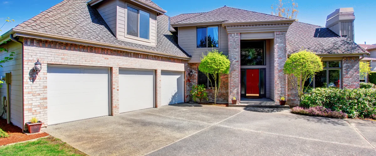 A luxurious brick house with a large paved driveway, three white garage doors, shrubs, and a bright red front door.
