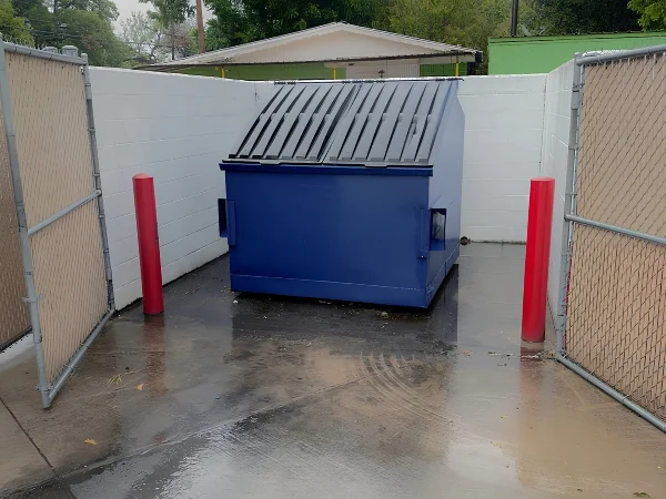 A blue dumpster with a ribbed lid sits within a white-walled, fenced alcove on wet concrete, flanked by red bollards.