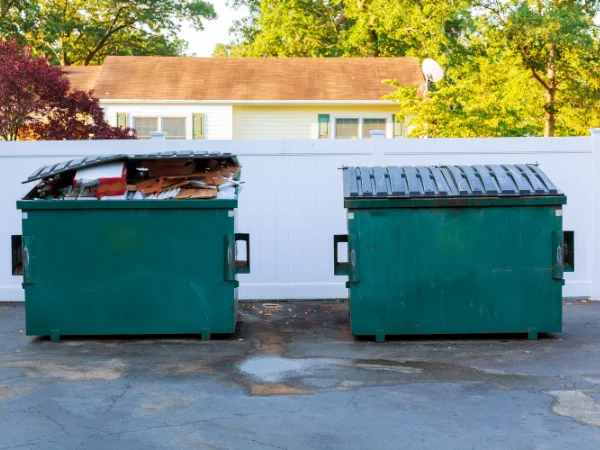 Two green dumpsters, one overflowing, in front of a white fence with a house and trees in the background.