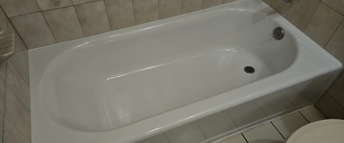 A sparkling clean white bathtub with a drain and overflow plate, set against light brown tiled walls.