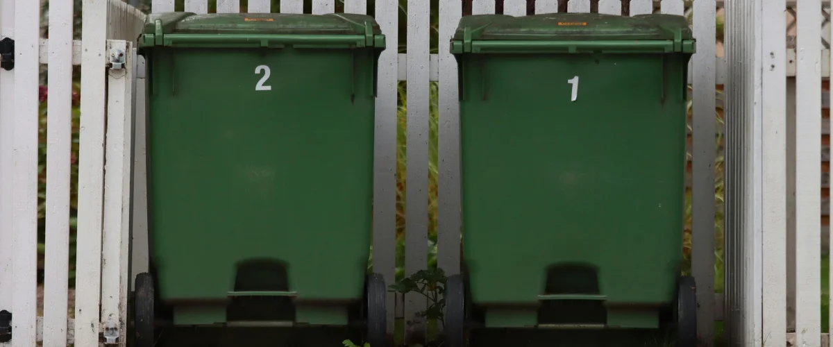 Two large green recycling bins with white recycling symbols sit on the edge of an asphalt road next to grass. Two large green recycling bins with white recycling symbols sit on the edge of an asphalt road next to grass.