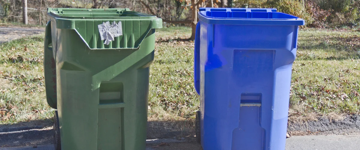 A green curbside trash can with a partial sticker and a blue recycling bin stand side-by-side on asphalt. A green curbside trash can with a partial sticker and a blue recycling bin stand side-by-side on asphalt.