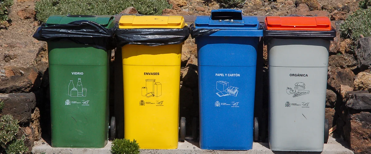 Four different colored recycling bins with Spanish labels for glass, containers, paper/cardboard, and organic waste.