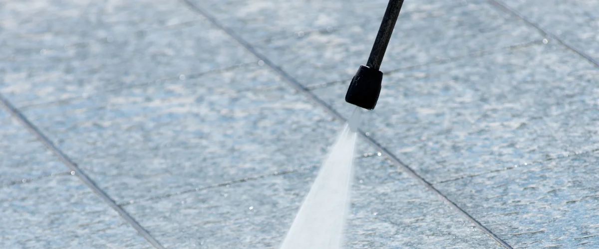 A close-up of a pressure washer nozzle spraying water onto light blue concrete patio tiles. A close-up of a pressure washer nozzle spraying water onto light blue concrete patio tiles.