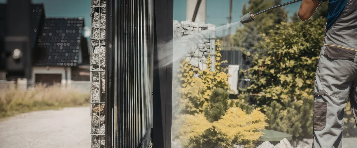 A man in work clothes using a pressure washer to clean a modern fence panel with a gabion wall section. A man in work clothes using a pressure washer to clean a modern fence panel with a gabion wall section.