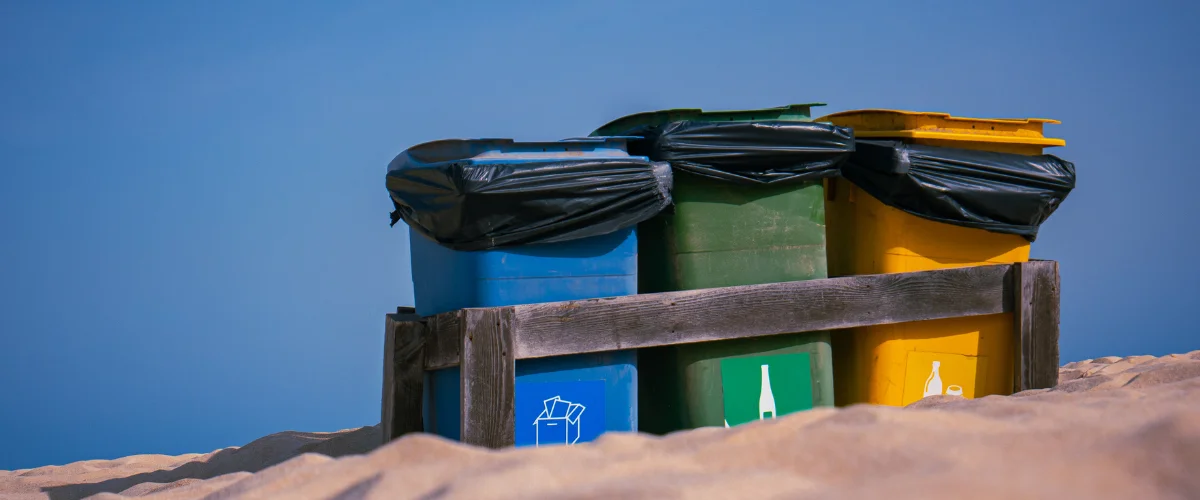 Three colorful recycling bins, blue, green, and yellow, with black bags seen against a blue sky on a sandy beach. Three colorful recycling bins, blue, green, and yellow, with black bags seen against a blue sky on a sandy beach.