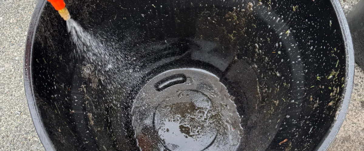A close-up, high-angle shot of a black garden pot being cleaned with a hose, showing water spraying inside.