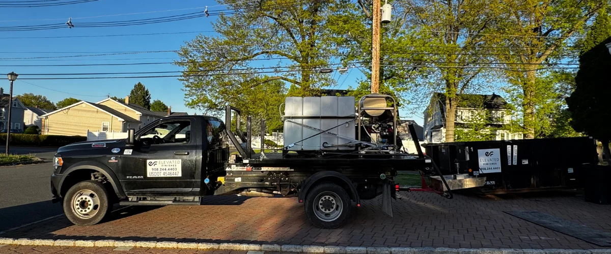 A black Elevated Finishes flatbed truck with a large tank and equipment parked on a brick sidewalk next to a street.