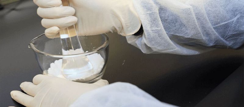 Gloved hands use a pestle to prepare white powder in a glass vessel.