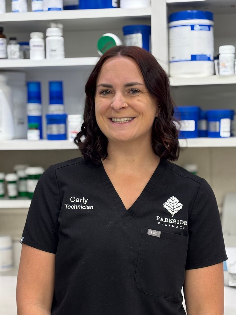 Carly smiles in black Parkside scrubs embroidered with her name and technician role, with preparation containers on the shelves behind her.