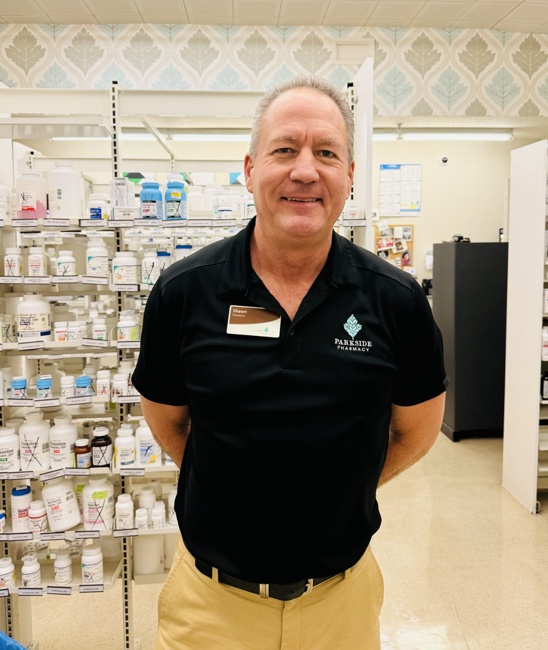 A smiling Parkside pharmacist in a branded black polo stands in front of stocked pharmacy shelves.