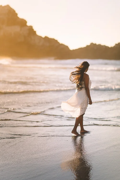 A woman in a white dress walking along the shoreline at sunset