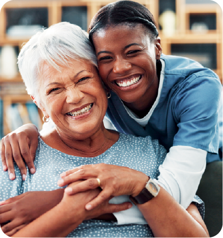 A dark nurse smiling and hugging a smiling white elderly woman from behind