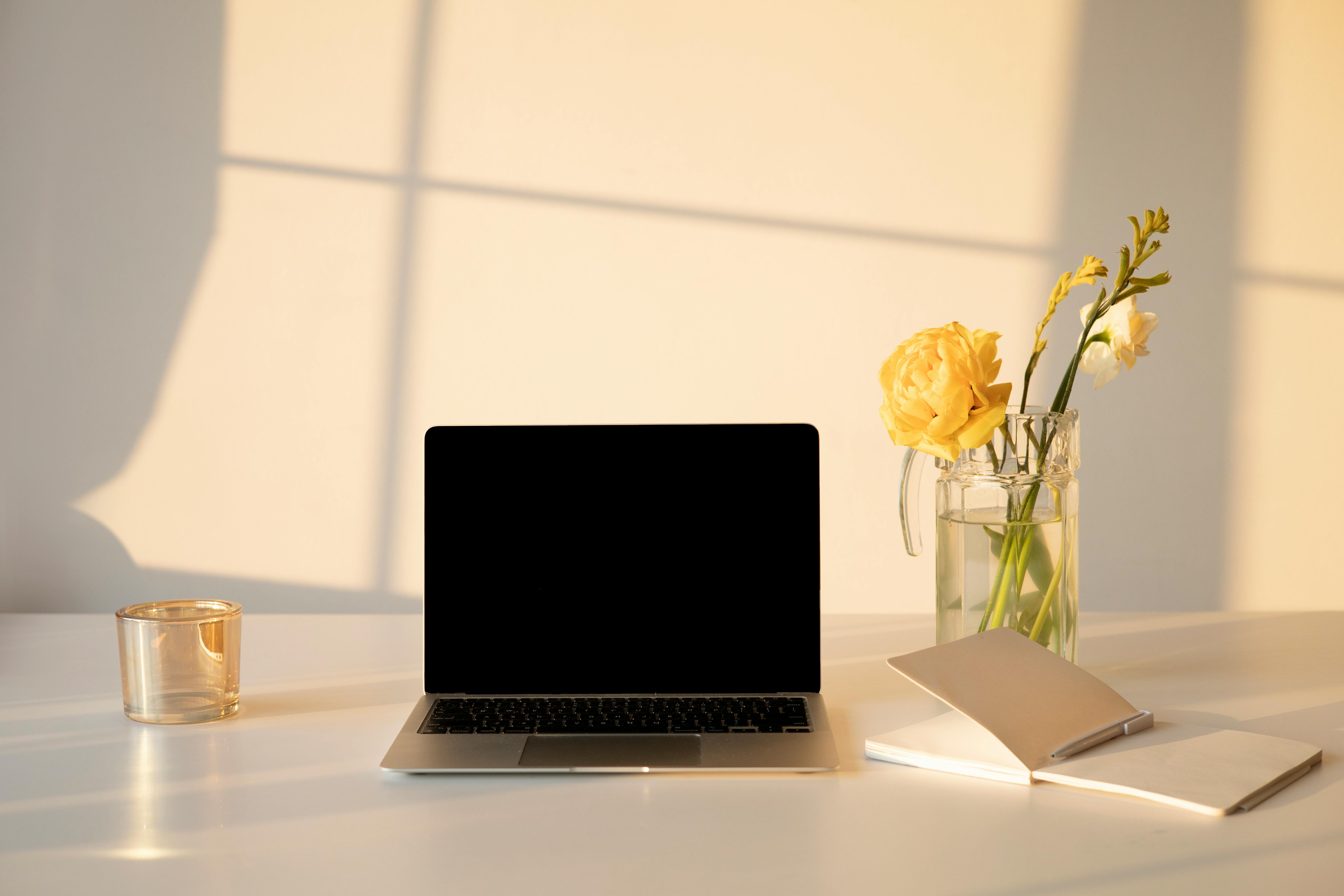 A laptop and open notebook on a clean desk in warm natural light, with a glass vase of yellow flowers.