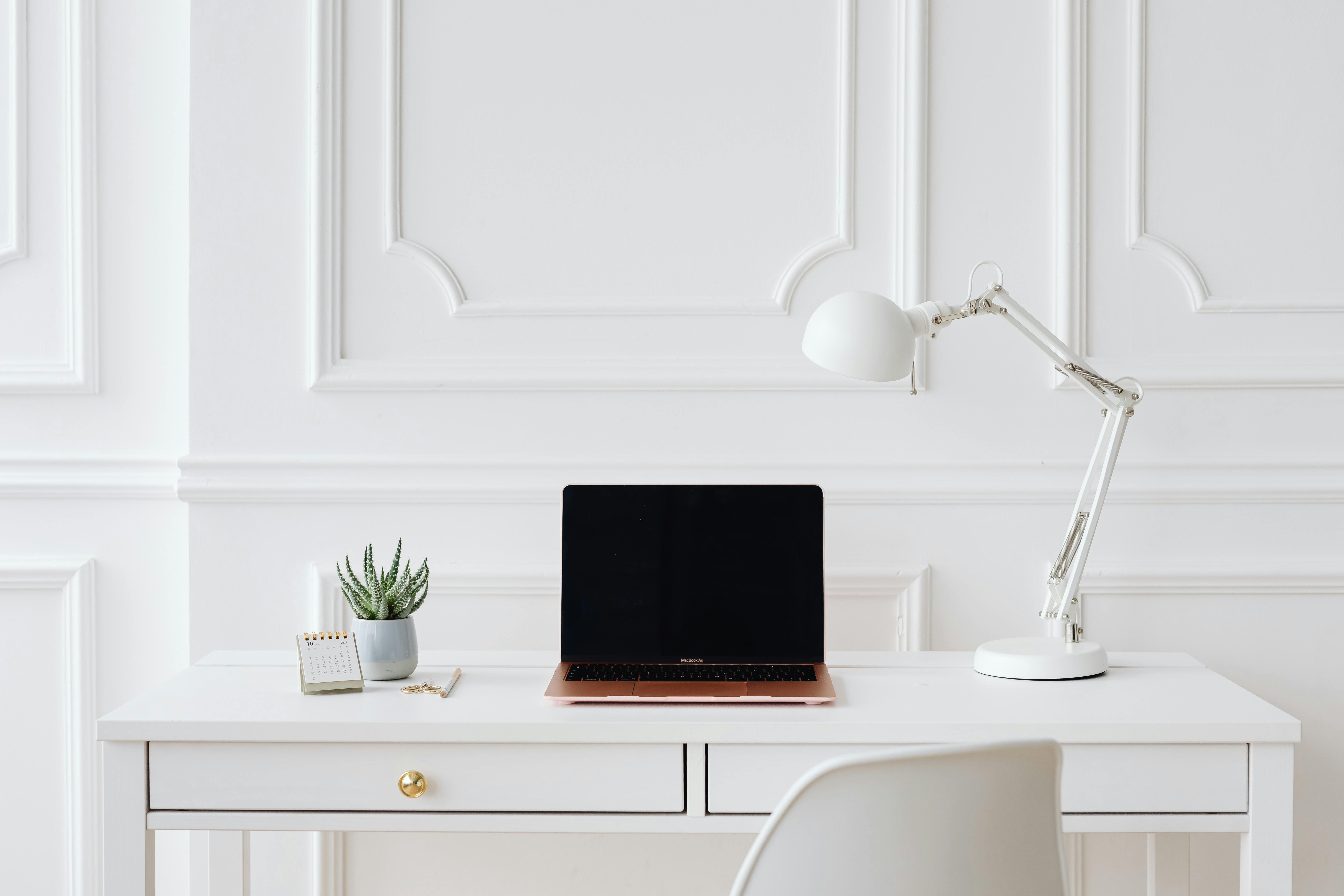 A clean white home office desk with an open laptop, arc lamp and small plant, representing a structured and considered approach to social media management for small business. A clean white home office desk with an open laptop, arc lamp and small plant, representing a structured and considered approach to social media management for small business.