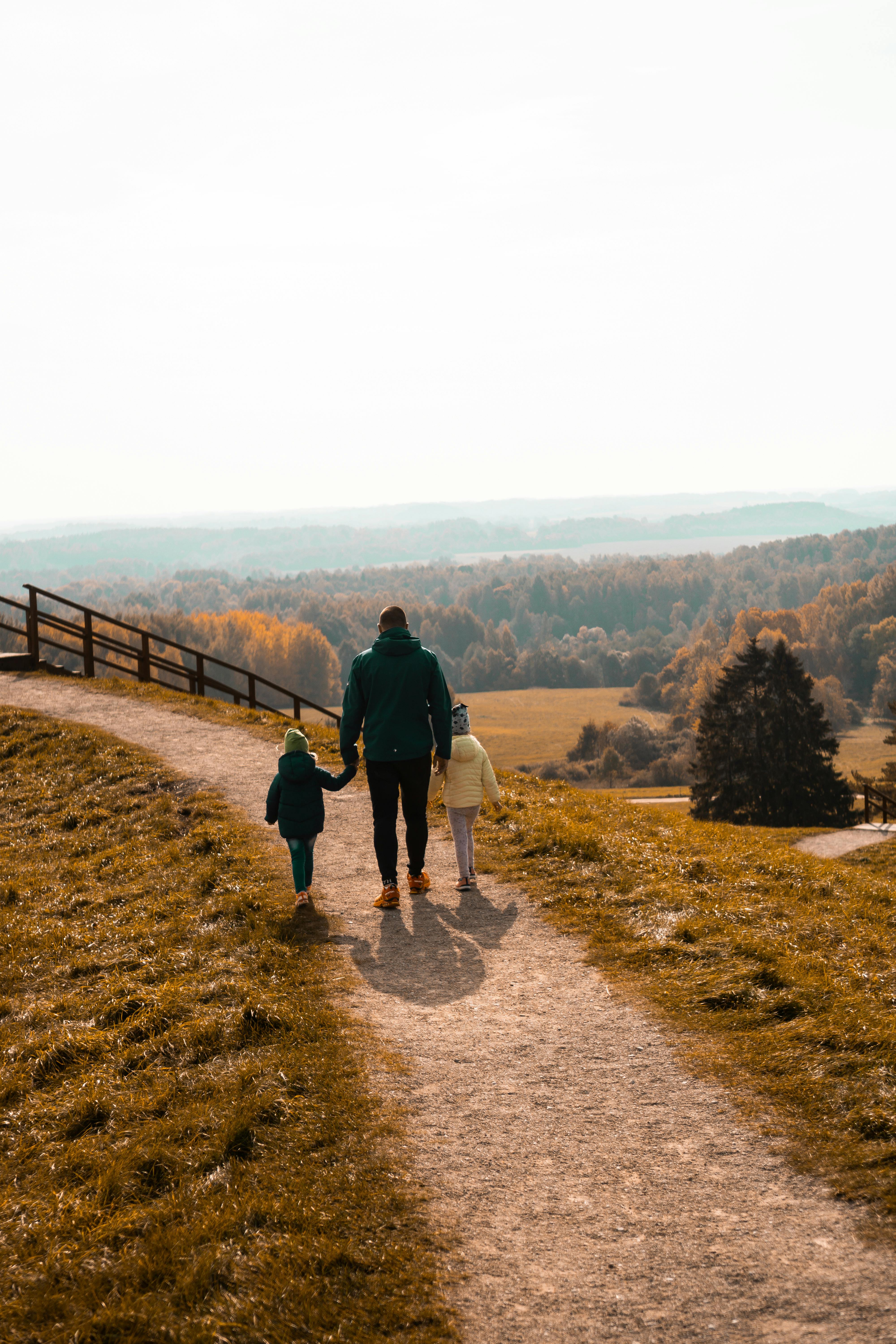 A parent walks along an open hillside path holding the hands of two young children, viewed from behind with a wide landscape ahead of them A parent walks along an open hillside path holding the hands of two young children, viewed from behind with a wide landscape ahead of them