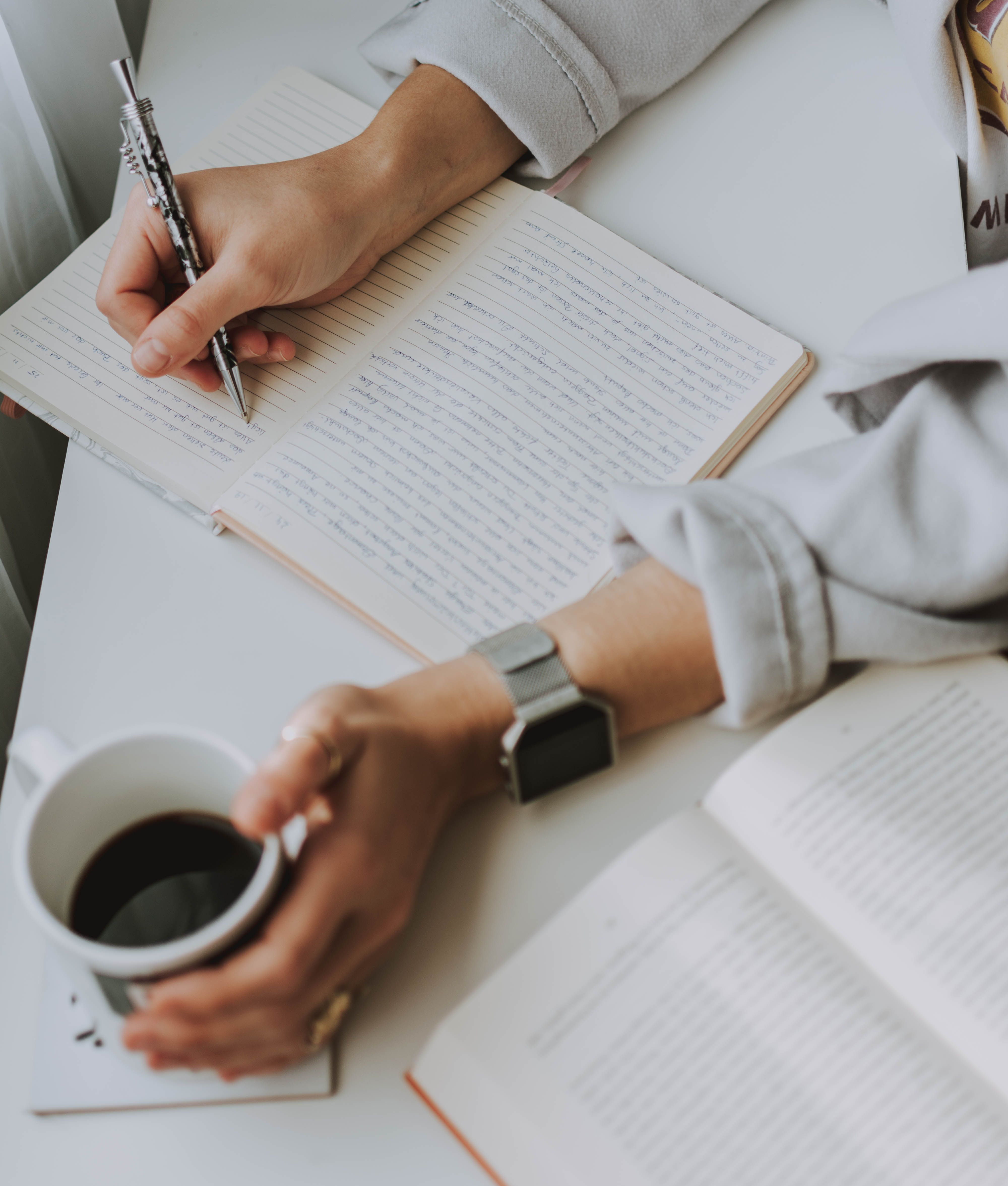 A woman writing in a notebook while holding a coffee cup, viewed from above, with a second open book alongside her. A woman writing in a notebook while holding a coffee cup, viewed from above, with a second open book alongside her.