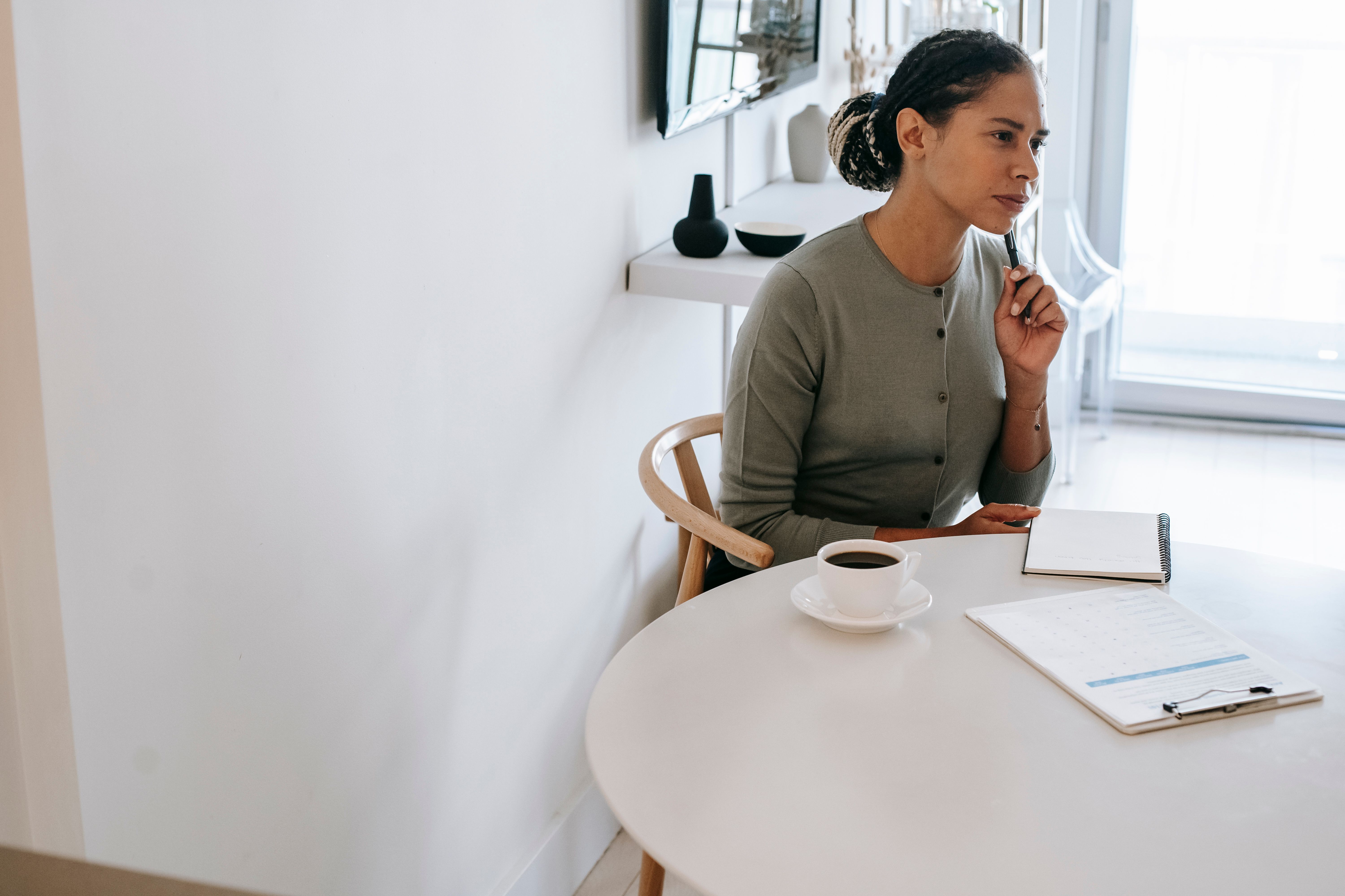 A business owner sitting at a table with a notebook and coffee looking thoughtfully to the side considering her customer relationships.