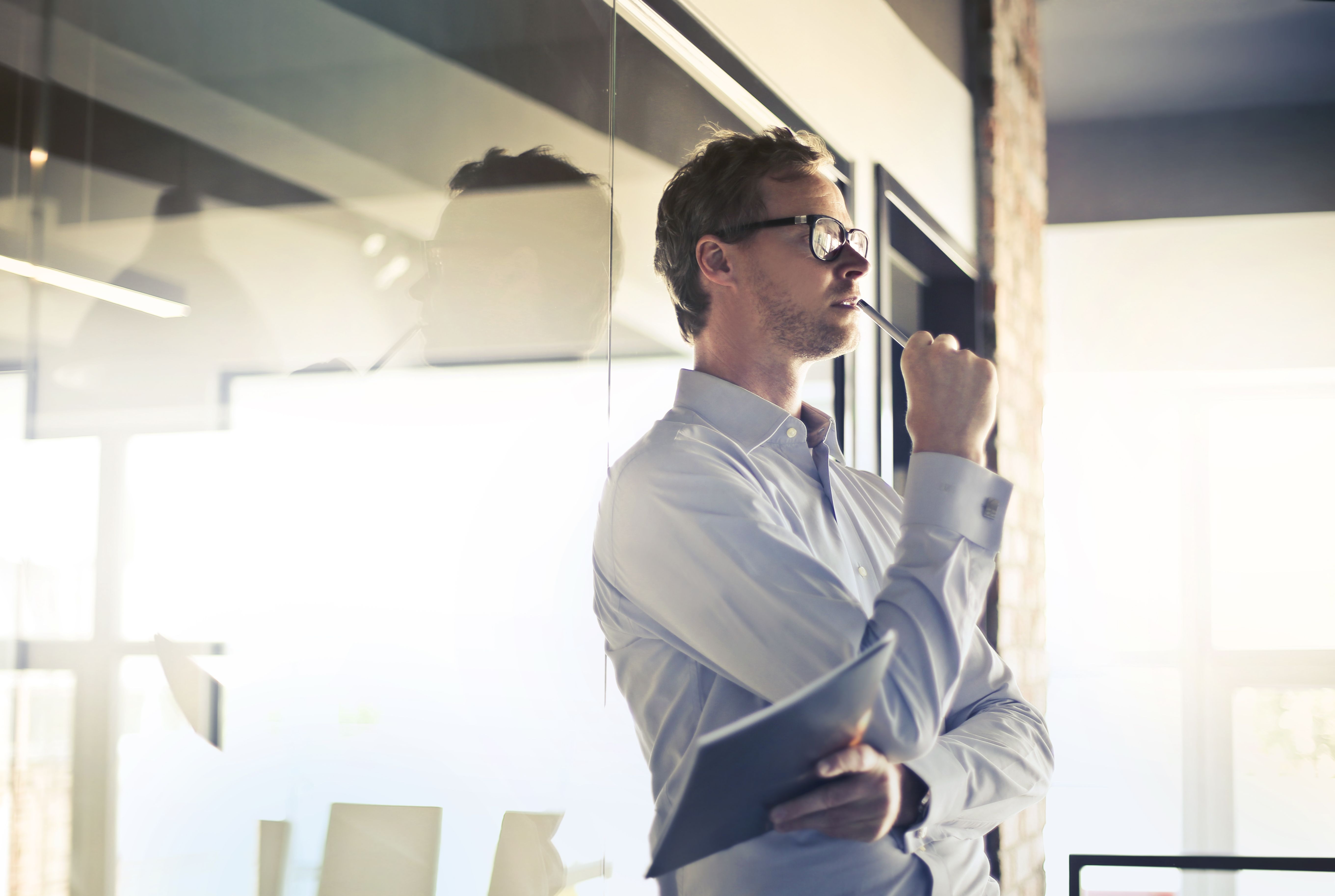 A male business owner standing in a bright office space holding a notebook and looking forward thoughtfully considering his next strategic decision.