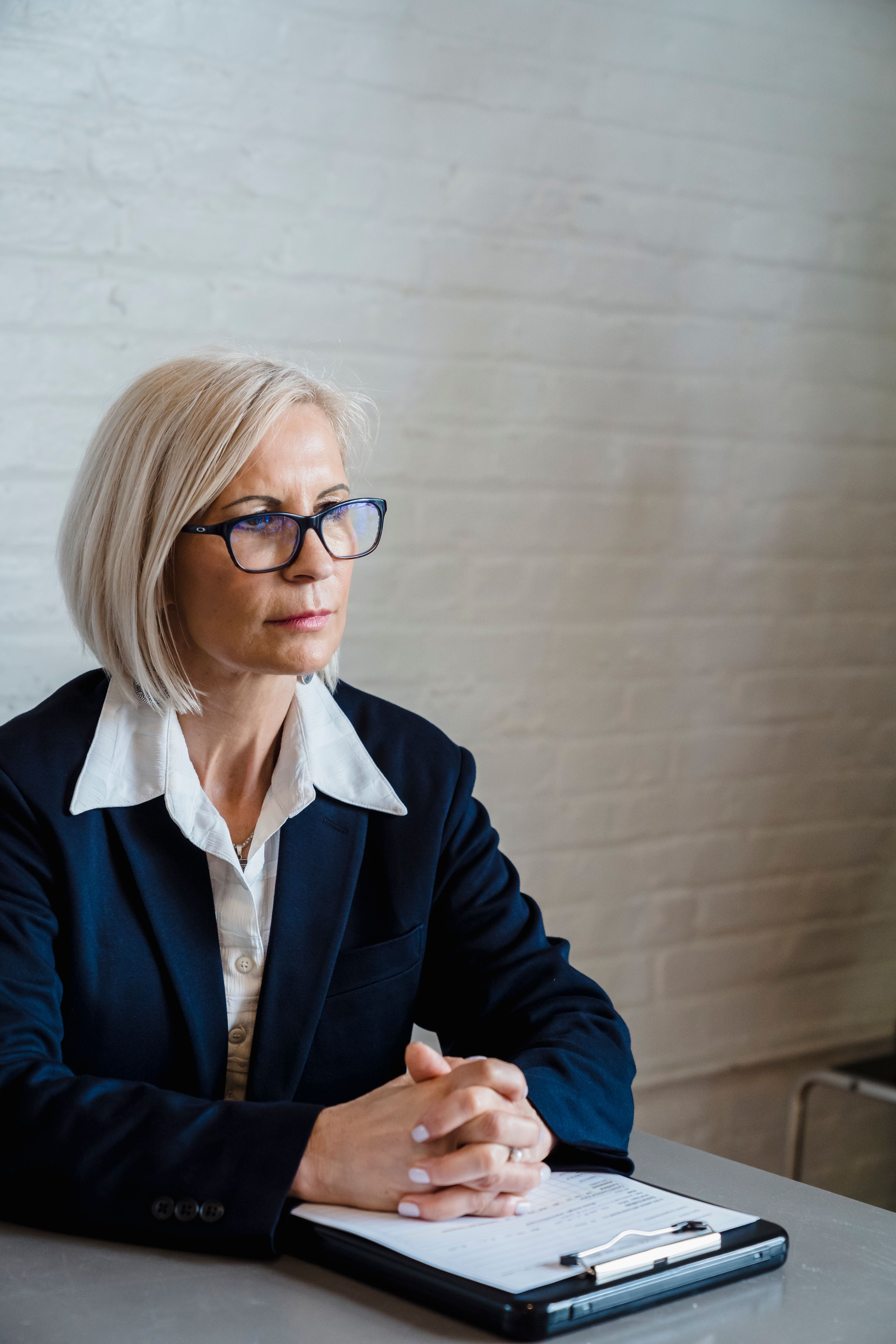 An established female business owner sitting at a table with a clipboard, hands folded, looking forward with quiet intensity as she considers where her business actually stands.