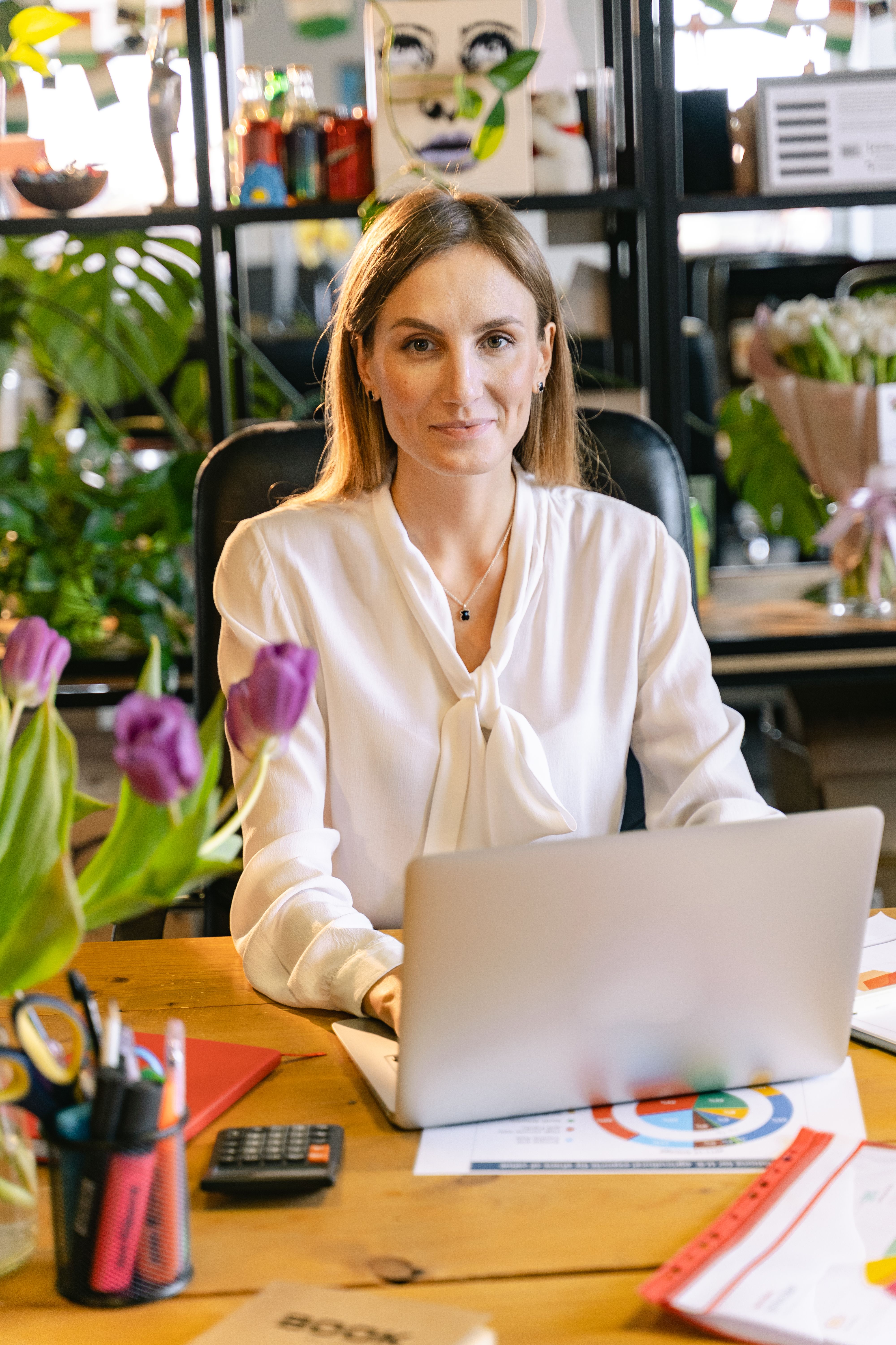 A business owner at her desk with a calm settled expression, surrounded by the warmth of a creative professional workspace, representing the trust built through consistent delivery that brings clients back without having to ask.