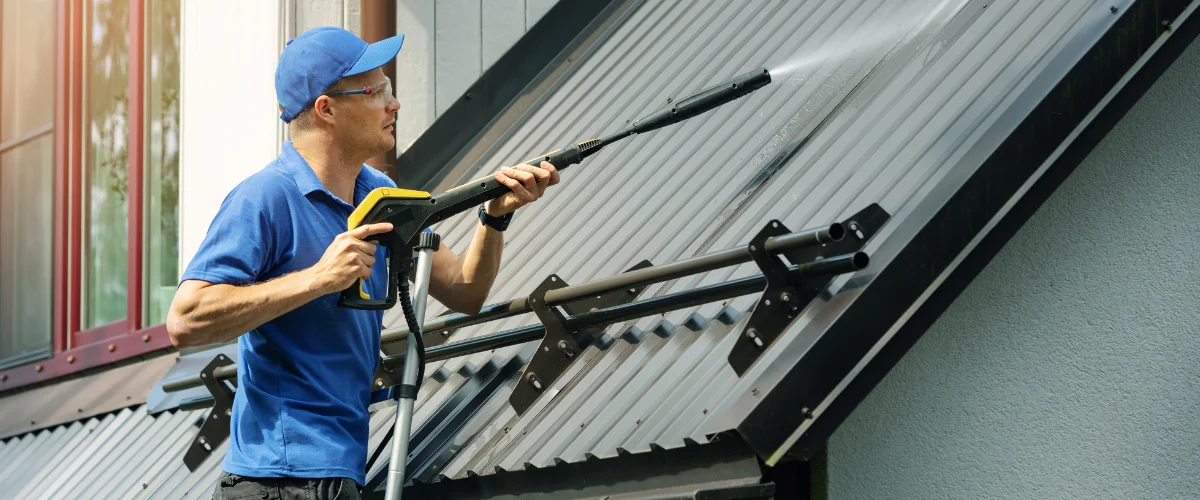 A man in a blue cap power washing a gray corrugated metal roof, removing dirt and grime. A man in a blue cap power washing a gray corrugated metal roof, removing dirt and grime.