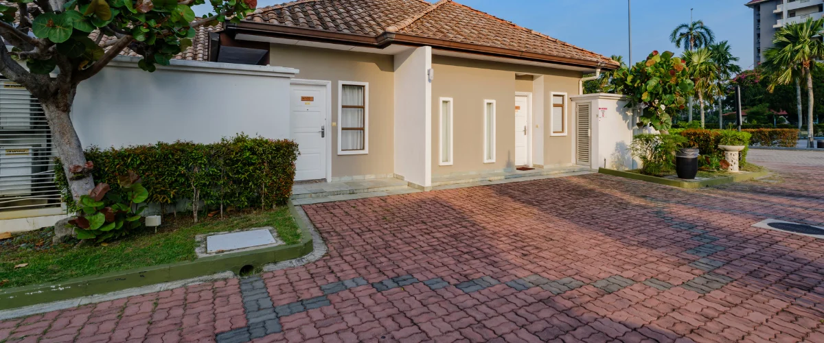 A tan house with a terracotta tile roof and red brick driveway, surrounded by lush green foliage and palm trees. A tan house with a terracotta tile roof and red brick driveway, surrounded by lush green foliage and palm trees.