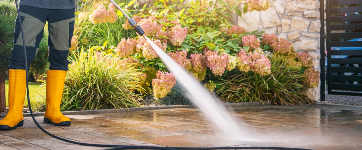 A person in yellow rubber boots power washing a patio made of paving stones next to flowering pink bushes. A person in yellow rubber boots power washing a patio made of paving stones next to flowering pink bushes.