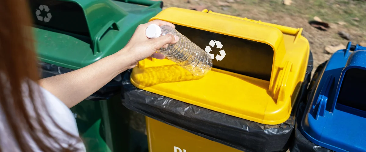 A person's hand placing an empty plastic water bottle into a yellow recycling bin with a recycle symbol. A person's hand placing an empty plastic water bottle into a yellow recycling bin with a recycle symbol.