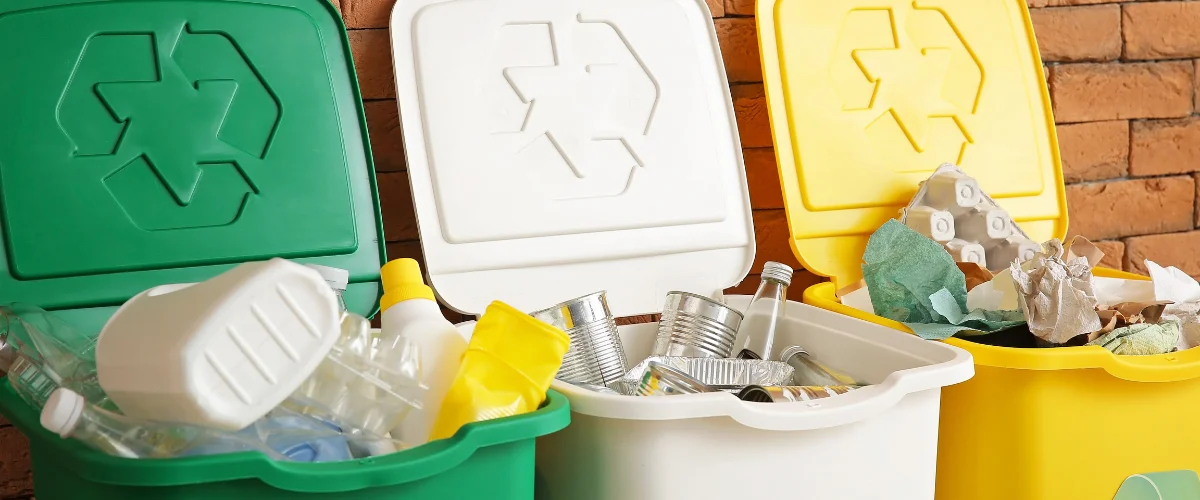 Three color-coded recycling bins, green for plastics, white for metals, and yellow for paper, full against a brick wall. Three color-coded recycling bins, green for plastics, white for metals, and yellow for paper, full against a brick wall.