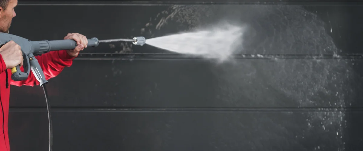 A man in a red shirt power washing a dirty gray garage door with a high-pressure spray. A man in a red shirt power washing a dirty gray garage door with a high-pressure spray.