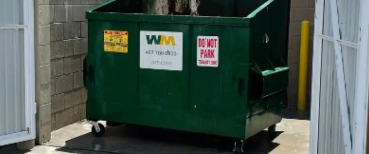 A large green Waste Management dumpster with yellow and red warning signs sits outdoors next to a block wall.
