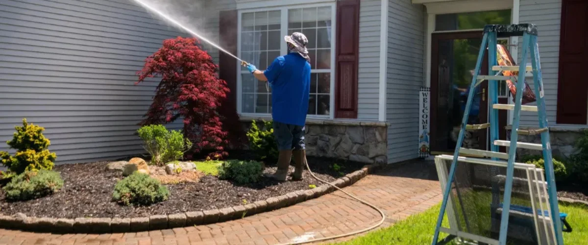 A person uses a pressure washer to clean dirty yellow patio pavers, revealing clean, wet black pavers beyond. A person uses a pressure washer to clean dirty yellow patio pavers, revealing clean, wet black pavers beyond.