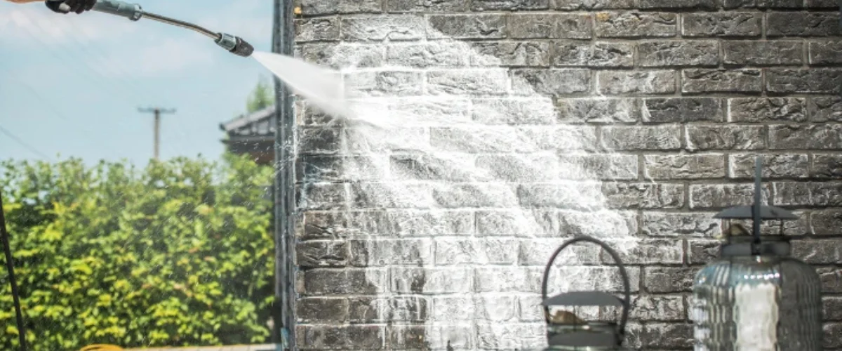 A pressure washer spraying water onto a dark brick wall, creating a clean streak, with green bushes in the background. A pressure washer spraying water onto a dark brick wall, creating a clean streak, with green bushes in the background.