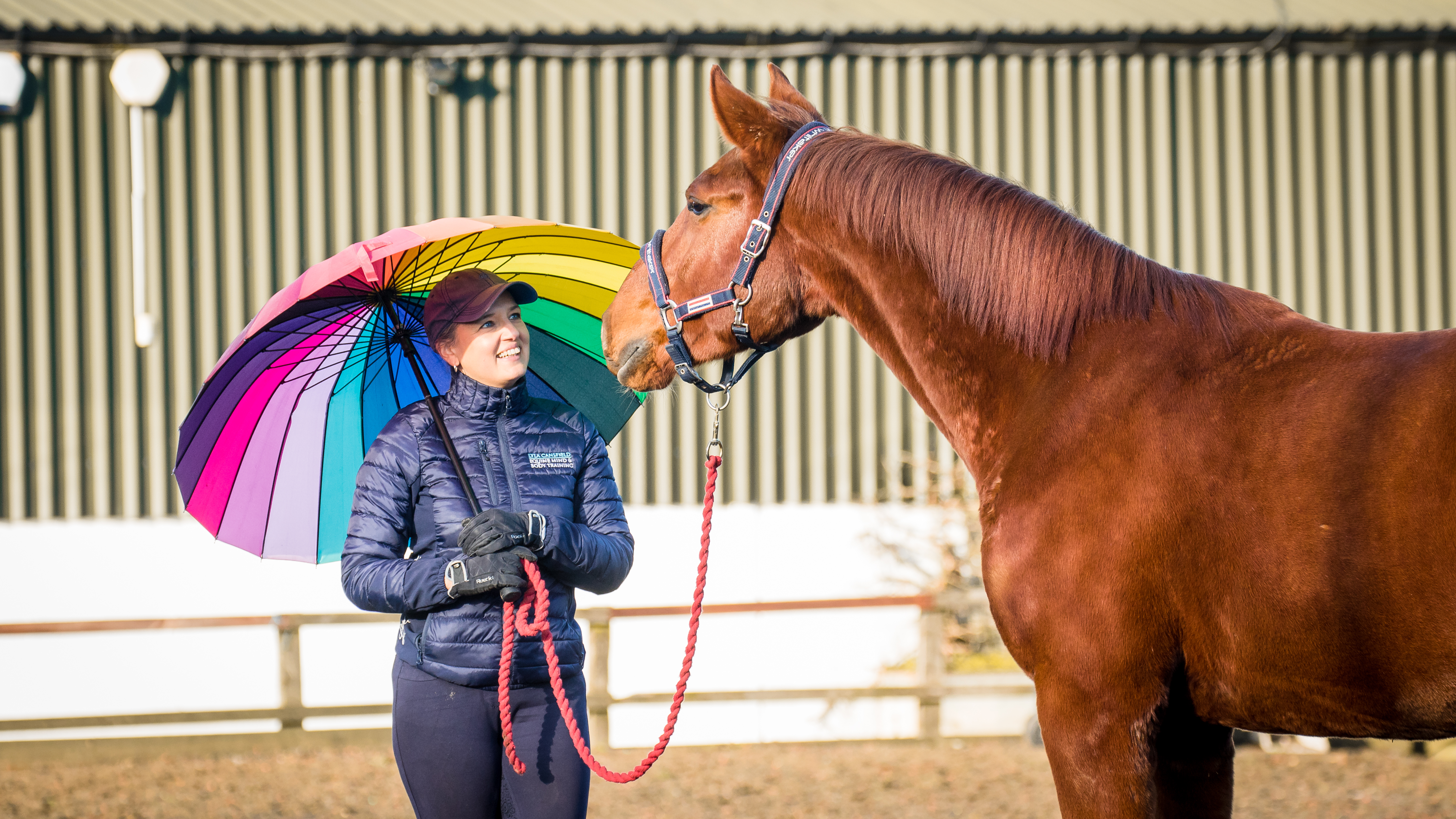 Lyla holding a rainbow umbrella beside a relaxed chestnut horse