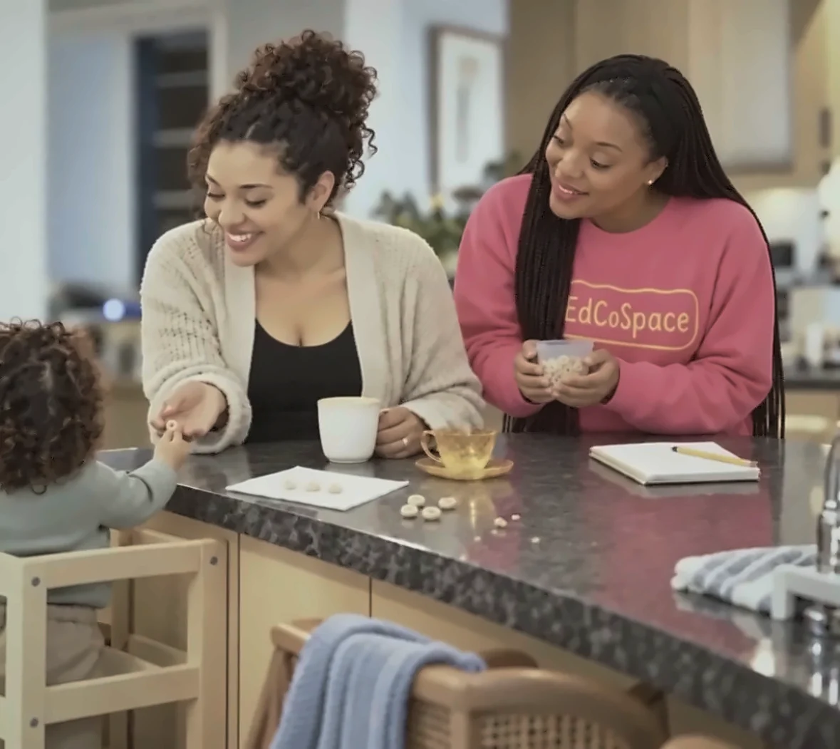 Two women smiling with a toddler on a learning tower at a kitchen counter