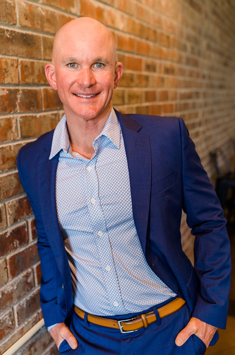 Allan Blain smiling in a sharp blue suit while standing against a brick wall, founder of Life’s Hard Succeed Anyway Podcast.