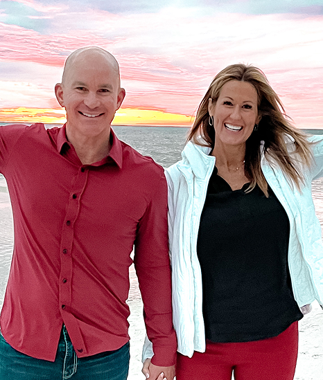 Allan and Nicole Blain hold hands on the beach at sunset, dressed in red and white, exuding joy and connection — representing their commitment to building a life of shared purpose and performance.