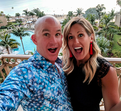 Allan and Nicole Blain take a joyful selfie on a tropical balcony with a resort view, palm trees, and pool in the background — showcasing their adventurous lifestyle.