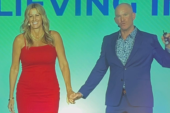 Allan and Nicole Blain on stage holding hands, smiling and speaking to an audience—representing unity, leadership, and inspirational coaching.