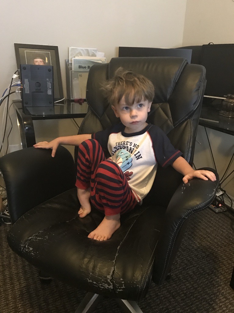 Young child sitting in a worn office chair in the home office where dad works from home