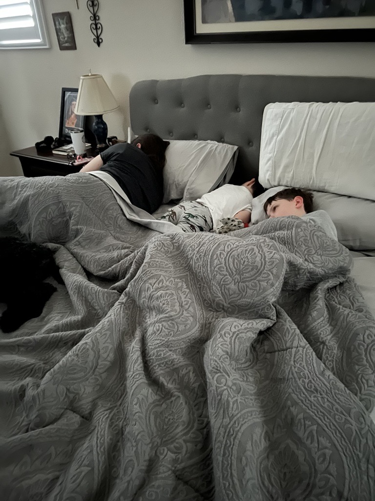 A tired parent and two children asleep together in the same bed under a gray comforter on a school morning.