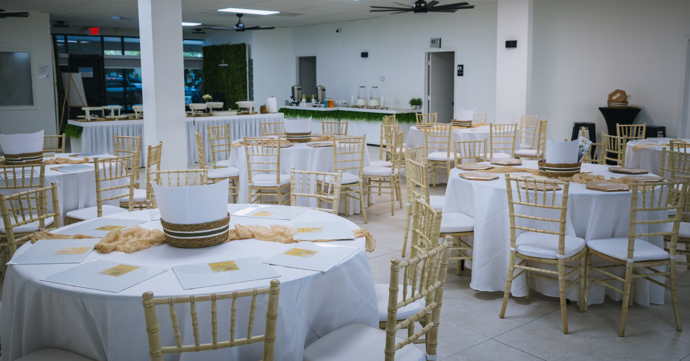 Elegant event setup with round tables, white linens, and gold chiavari chairs at a Lauderdale Lakes event space with catering area and modern decor