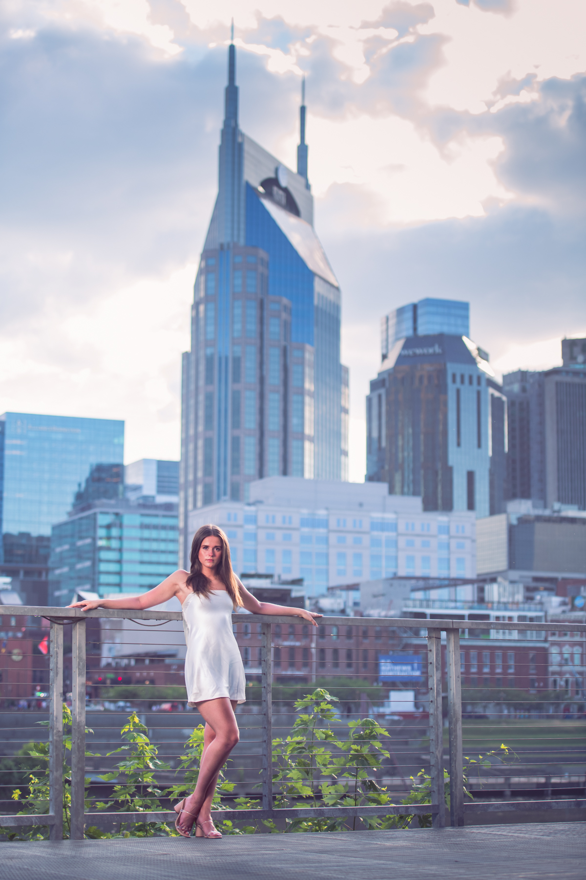 A high school senior in a white satin slip dress leans against the railing at Wasioto Park, arms resting wide and gaze steady, as the Nashville skyline and the AT&T Building rise behind her across the Cumberland River under soft evening clouds. A poised, city-ready Nashville skyline senior portrait by Cate Connery Bury Photography A high school senior in a white satin slip dress leans against the railing at Wasioto Park, arms resting wide and gaze steady, as the Nashville skyline and the AT&T Building rise behind her across the Cumberland River under soft evening clouds. A poised, city-ready Nashville skyline senior portrait by Cate Connery Bury Photography