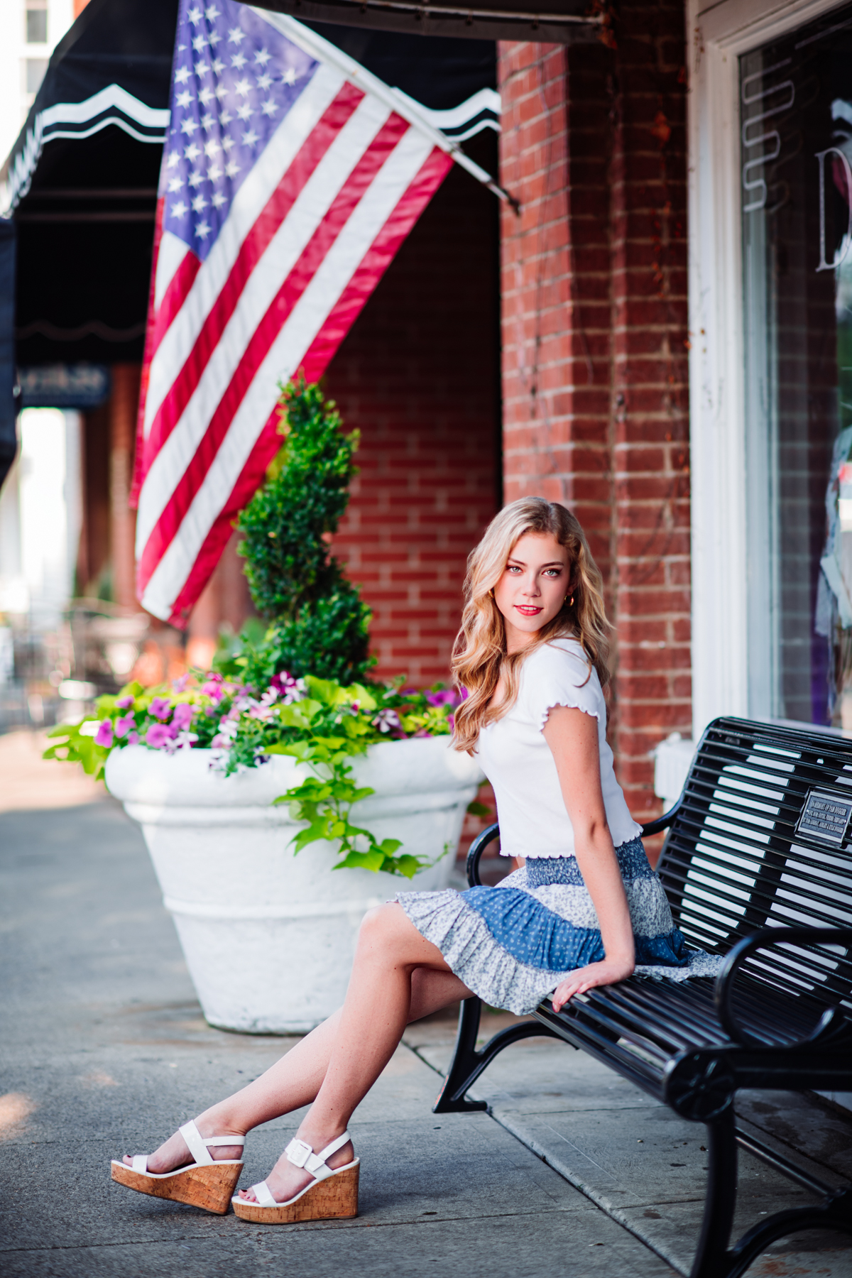 A high school senior in a white tee and blue tiered floral skirt sits on a black bench along Main Street in Downtown Franklin, glancing back over her shoulder with quiet confidence, an American flag, brick storefront, and planter of pink petunias framing her. A classic Franklin, Tennessee senior portrait by Cate Connery Bury Photography, full of hometown charm. A high school senior in a white tee and blue tiered floral skirt sits on a black bench along Main Street in Downtown Franklin, glancing back over her shoulder with quiet confidence, an American flag, brick storefront, and planter of pink petunias framing her. A classic Franklin, Tennessee senior portrait by Cate Connery Bury Photography, full of hometown charm.
