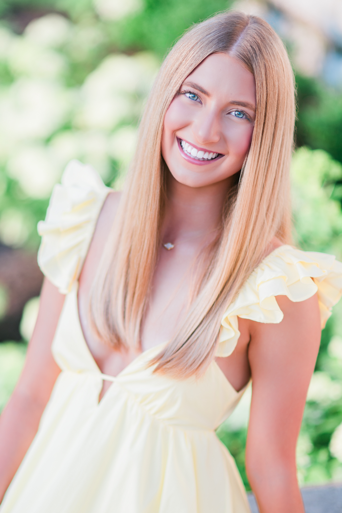 A young woman with long blonde hair smiles warmly at the camera while wearing a soft yellow dress with ruffled sleeves. Her bright blue eyes catch the light, and a delicate necklace rests at her collarbone. Behind her, creamy white blooms and lush greenery blur into a dreamy backdrop, creating a portrait filled with light and softness.  She leans slightly forward, relaxed and confident, as if sharing a joyful conversation with a close friend. The gentle yellow of her dress echoes the warmth of a summer afternoon, while her genuine smile radiates kindness and optimism. The close composition draws attention to her expression, allowing her personality to become the heart of the image.  This portrait celebrates youthful confidence, beauty, and authenticity. The soft colors, glowing light, and natural surroundings create a feeling of hope and possibility, capturing a moment that feels both timeless and full of promise.