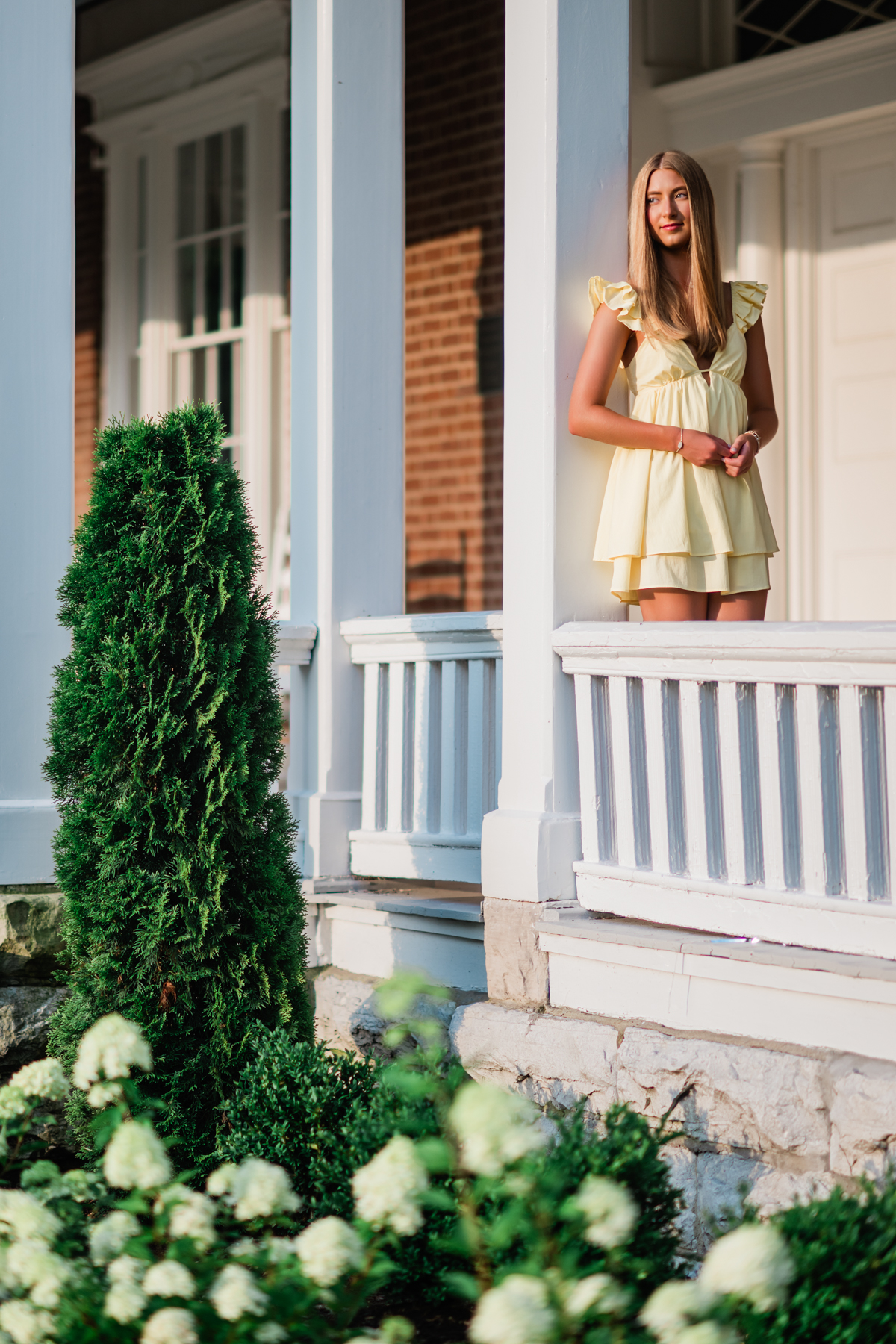 A young woman with long blonde hair looks off in the distance while wearing a soft yellow dress with ruffled sleeves. Her bright blue eyes catch the light, and a delicate necklace rests at her collarbone. She stands on the porch of a beautiful historical home, creating a portrait filled with light and softness.  She leans slightly forward, relaxed and confident, as if sharing a joyful conversation with a close friend. The gentle yellow of her dress echoes the warmth of a summer afternoon, while her genuine smile radiates kindness and optimism. The close composition draws attention to her expression, allowing her personality to become the heart of the image.  This portrait celebrates youthful confidence, beauty, and authenticity. The soft colors, glowing light, and natural surroundings create a feeling of hope and possibility, capturing a moment that feels both timeless and full of promise.
